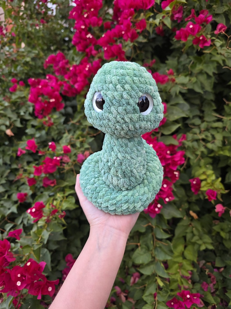 Pu&ograve; includere: Un giocattolo a forma di serpente di peluche verde chiaro fatto a mano con grandi occhi con pupille nere. Il morbido serpente strutturato &egrave; arrotolato e tenuto contro uno sfondo di vibranti fiori rosa e fogliame verde.