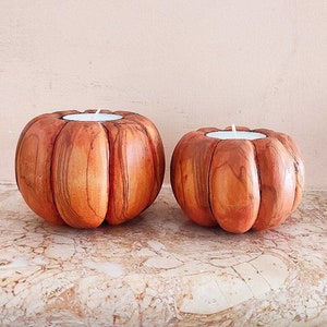Può includere: Due portacandele in legno a forma di zucca con candele tealight bianche all'interno. I portacandele sono realizzati in legno naturale di colore marrone e arancione.