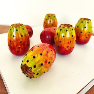 Könnte beinhalten: Sechs handbemalte Keramik-Ornamente in Form von Kaktusfeigenfrüchten in Rot-, Orange- und Gelbtönen mit schwarzen Punkten. Die Ornamente sind auf einer weißen Oberfläche angeordnet.