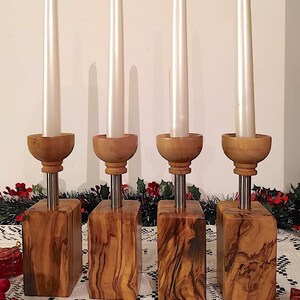 Può includere: Set di quattro portacandele in legno con finitura in legno naturale. Ogni portacandele presenta una piccola ciotola nella parte superiore e un'asta di metallo che si estende al centro. I portacandele sono disposti in fila su una tovaglia bianca.