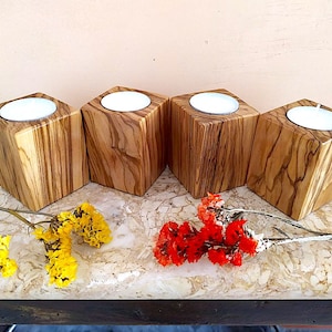 Può includere: Quattro portacandele in legno con finitura a grana di legno naturale. Ogni portacandele contiene una piccola candela rotonda bianca. I portacandele sono disposti in fila su una superficie di colore chiaro con fiori gialli e rossi in primo piano.
