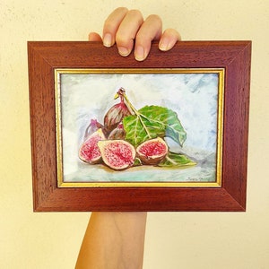 K&ouml;nnte beinhalten: Ein gerahmtes Gem&auml;lde eines Stilllebens mit einer Gruppe reifer Feigen und einem gr&uuml;nen Blatt. Die Feigen sind in Schattierungen von Lila, Rosa und Gr&uuml;n gemalt, und das Blatt ist ein leuchtendes Gr&uuml;n. Das Gem&auml;lde ist vor einem hellblauen Hintergrund platziert und in einem dunklen Holzrahmen gerahmt.