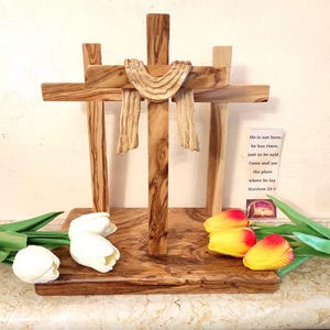 Può includere: Una scultura a forma di croce in legno con tre croci, un drappo e una base. La scultura è realizzata in legno marrone chiaro con venature naturali. Davanti ci sono tulipani bianchi e fiori gialli e rossi. È visibile una scheda con testo.