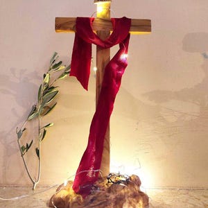 Può includere: Una croce di legno con un panno rosso drappeggiato sopra, illuminata da piccole luci fatate. La croce ha le lettere "INRI" in alto. La base è in legno naturale strutturato. Un rametto di verde è a sinistra.