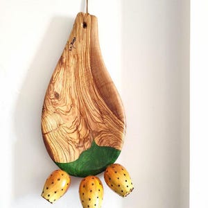 May include: A teardrop-shaped olive wood chopping board with a green epoxy resin inlay and three small yellow prickly pear fruits hanging from the bottom.