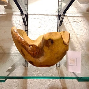 May include: A large, natural wood bowl with a unique, organic shape. The bowl is light brown with darker brown streaks and knots. It is displayed on a glass shelf with a white background.