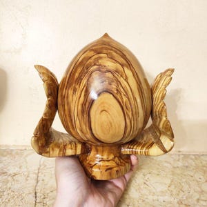 May include: A handcrafted wooden sculpture with a bulbous, egg-like form and wing-like extensions. The sculpture is made of light brown wood with darker brown grain patterns, and it sits on a small pedestal.