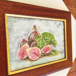 K&ouml;nnte beinhalten: Ein gerahmtes Gem&auml;lde mit einem Stillleben aus Feigen und einem Blatt. Die Feigen sind in Schattierungen von Lila, Rosa und Gr&uuml;n gemalt. Das Blatt ist dunkelgr&uuml;n. Das Gem&auml;lde ist in einem dunklen Holzrahmen mit goldener Verzierung gerahmt.