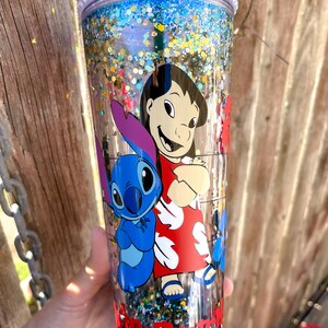 May include: A clear tumbler with a blue and gold glitter design. The tumbler features a cartoon image of Lilo and Stitch from the Disney movie Lilo & Stitch. The tumbler has a clear lid with a silver glitter rim.