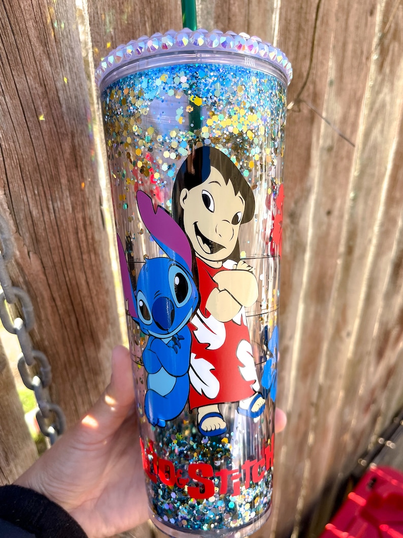 May include: A clear tumbler with a blue and gold glitter design. The tumbler features a cartoon image of Lilo and Stitch from the Disney movie Lilo & Stitch. The tumbler has a clear lid with a silver glitter rim.