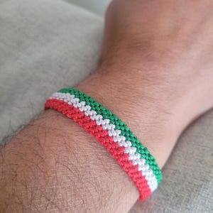 Puede incluir: Una pulsera hecha a mano con un patrón tejido en rojo, blanco y verde. La pulsera se lleva en la muñeca y parece estar hecha de un material suave y flexible. Los colores están dispuestos en franjas horizontales.