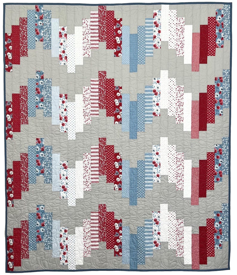 May include: A patchwork quilt with a grey background and a repeating pattern of red, white, and blue rectangular blocks. The blocks feature floral, polka dot, and striped patterns. The quilt is edged with a dark blue binding.