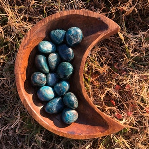 May include: A collection of blue-green apatite tumbled stones in a wooden crescent moon-shaped bowl. The stones are smooth and polished, with a variety of shapes and sizes.