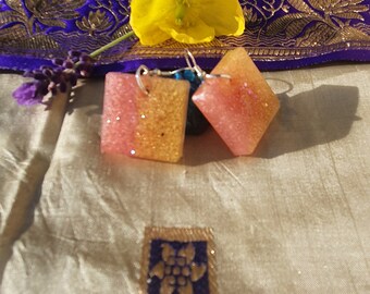 May include: A pair of square-shaped earrings with pink and gold glitter embedded in the resin. The earrings have silver hooks.