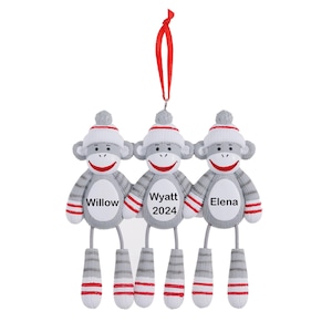May include: Three gray sock monkey ornaments with red accents. The monkeys are holding hands and have personalized names: Willow, Wyatt 2024, and Elena.