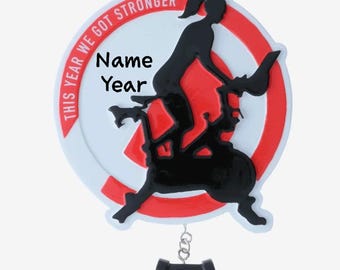 Op de afbeelding: Witte en rode ronde ornament met een zwart silhouet van een persoon die op een hometrainer fietst. De ornament heeft de tekst "This year we got stronger" en een ruimte voor een naam en een jaar.