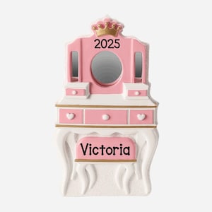May include: A pink and white vanity ornament with a mirror, drawers, and a stool. The ornament has a gold crown on top and the year "2025" is printed on the mirror. The name "Victoria" is printed on the stool.