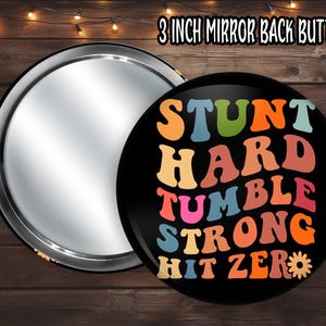 May include: A 3-inch mirror-backed button with a black face featuring the words "STUNT HARD TUMBLE STRONG HIT ZERO" in colorful, retro-style lettering. The mirror reflects light, and the button has a shiny silver edge.