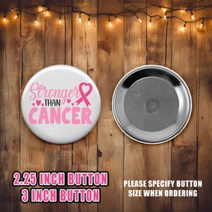 Op de afbeelding: Een witte knop met de tekst "Stronger Than Cancer" in roze letters en een roze lint. De knop is verkrijgbaar in de maten 2,25 inch en 3 inch. Geef de maat aan bij het bestellen.