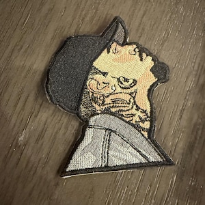 May include: Embroidered patch featuring a stylized profile of a person with a tattoo. The patch has a black hood, tan skin, and a gray jacket. The design is detailed with embroidered lines and textures, creating a unique and artistic look.
