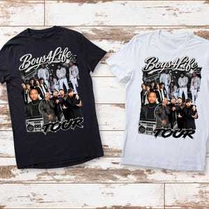 May include: Two t-shirts, one black and one white, featuring a graphic design. The design includes the text "Boys4Life" above a group of people and the word "TOUR" below. The shirts are displayed on a light brown wooden surface.