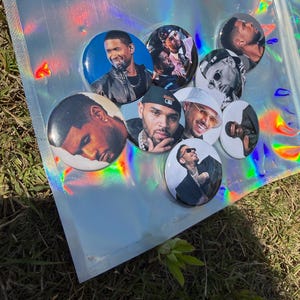 Può includere: Una collezione di bottoni rotondi con immagini di varie celebrità maschili. I bottoni sono disposti su una busta olografica iridescente, su uno sfondo di erba verde. I bottoni variano per dimensioni e design.