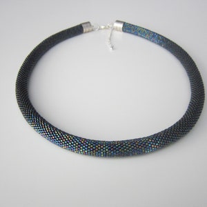 Op de afbeelding: Een zwarte kralenketting met iriserende accenten. De ketting is gemaakt van kleine, ronde kralen die in een strak patroon aan elkaar zijn geweven. De kralen zijn zwart met een subtiele iriserende glans die ze een licht regenboogachtig uiterlijk geeft.