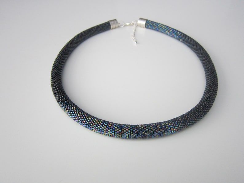Op de afbeelding: Een zwarte kralenketting met iriserende accenten. De ketting is gemaakt van kleine, ronde kralen die in een strak patroon aan elkaar zijn geweven. De kralen zijn zwart met een subtiele iriserende glans die ze een licht regenboogachtig uiterlijk geeft.