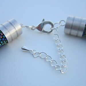 Op de afbeelding: Een close-up van een zilveren ketting met een sluiting en twee zilveren eindkappen. De ketting is gemaakt van kleine, iriserende kralen die een glinsterend effect cre&euml;ren.