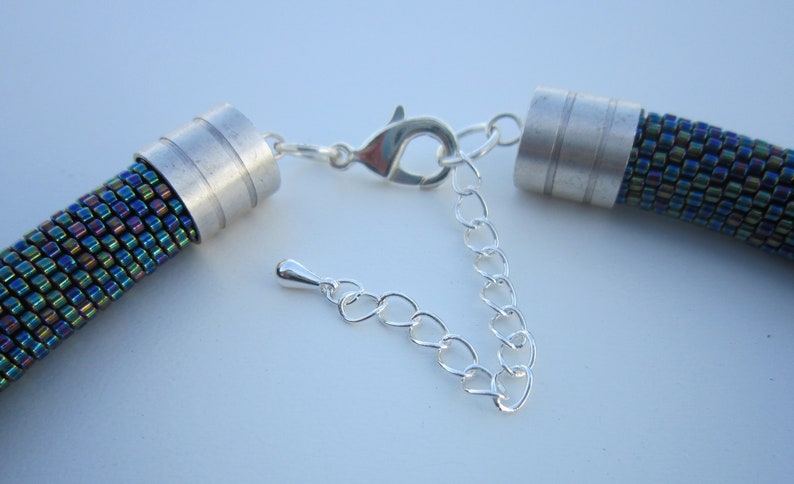 Op de afbeelding: Een close-up van een zilveren ketting met een sluiting en twee zilveren eindkappen. De ketting is gemaakt van kleine, iriserende kralen die een glinsterend effect cre&euml;ren.