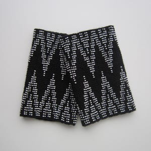 May include: Black and white beaded wrist cuffs with a geometric pattern. The cuffs are made of black fabric with white beads sewn into a chevron pattern.