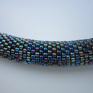 Op de afbeelding: Een close-up van een veelkleurige, iriserende kralenketting. De kralen zijn klein en rond en zijn gerangschikt in een strak, gelijkmatig patroon. De ketting is diepblauw met hints van groen, paars en goud.
