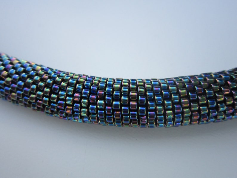 Op de afbeelding: Een close-up van een veelkleurige, iriserende kralenketting. De kralen zijn klein en rond en zijn gerangschikt in een strak, gelijkmatig patroon. De ketting is diepblauw met hints van groen, paars en goud.