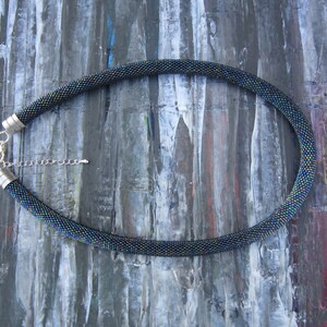 Op de afbeelding: Een zwarte kralenketting met iriserende kralen en zilveren sluitingen. De ketting ligt op een getextureerde, grijze achtergrond.