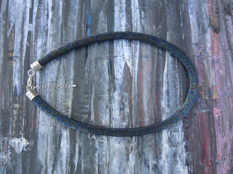Op de afbeelding: Een zwarte kralenketting met iriserende kralen en zilveren sluitingen. De ketting ligt op een getextureerde, grijze achtergrond.
