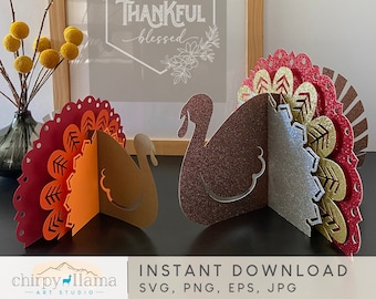 Può includere: Due tacchini di carta con accenti glitter. I tacchini sono rossi, arancioni, dorati e argentati. I tacchini sono in piedi su un tavolo. Il testo "GRATEFUL THANKFUL blessed" è su un cartello incorniciato dietro i tacchini.