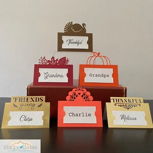 May include: Set of ten Thanksgiving place cards with various fall-themed designs. The cards are made of paper and feature a variety of colors, including red, orange, gold, and brown. The cards are decorated with turkeys, pumpkins, and leaves. The names "Clara", "Charlie", "Melissa", "Grandma", "Grandpa", and "Thankful" are written on the cards.