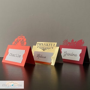 May include: Three red and gold place card holders with cut-out designs. The place cards read "Charlie", "Melissa", and "Grandma". One place card holder features a turkey design, another features the word "Thankful", and the third features a leaf design.