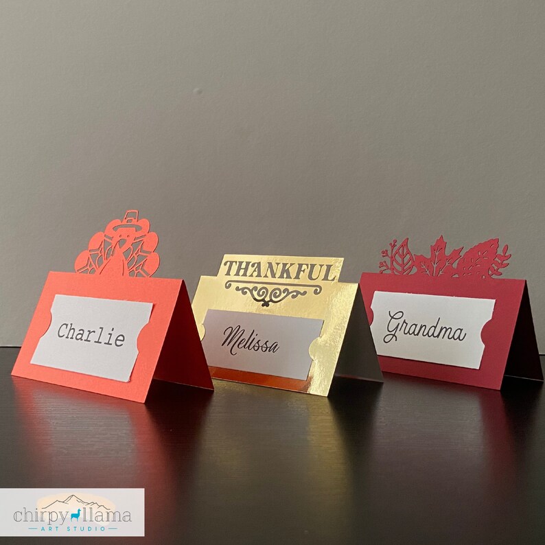 May include: Three red and gold place card holders with cut-out designs. The place cards read "Charlie", "Melissa", and "Grandma". One place card holder features a turkey design, another features the word "Thankful", and the third features a leaf design.