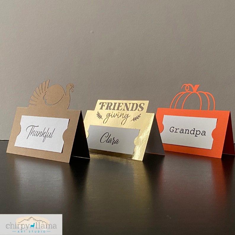May include: Three Thanksgiving place card holders. The first is brown with a turkey cutout and the text "Thankful". The second is gold with the text "FRIENDS giving" and "Clara". The third is orange with a pumpkin cutout and the text "Grandpa".