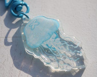 May include: A clear acrylic keyring with a light blue jellyfish design and glitter details. The keyring has a blue star-shaped clasp and a round ring. The jellyfish has a light blue head and white tentacles.