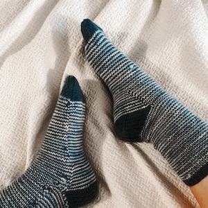 May include: A pair of knitted socks with a chevron pattern in shades of blue, white, and dark teal. The socks are on a beige textured surface. The heels and toes of the socks are a solid dark teal color.
