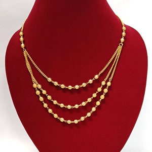 Könnte beinhalten: Eine mehrreihige Goldkette mit drei kaskadierenden Lagen aus feinen Goldketten. Jede Kette ist mit kleinen, gleichmäßig verteilten, gerippten Goldperlen besetzt, die eine strukturierte und reflektierende Oberfläche auf dem gesamten Schmuckstück erzeugen.