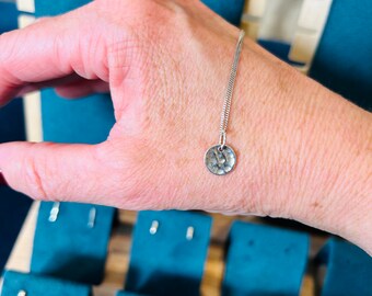May include: A silver necklace with a hammered round pendant. The necklace is on a person's wrist.