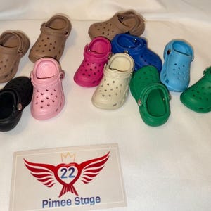 May include: A collection of ten miniature colorful clogs in various colors including black, pink, green, blue, and beige. The clogs are arranged on a white surface. The text "Pimee Stage" and a logo with a crown and wings are visible in the bottom right corner of the image.