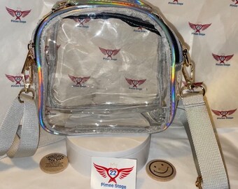 May include: A clear plastic crossbody bag with a silver iridescent trim and a gold chain strap. The bag has a zipper closure and a small pocket on the front. The bag is sitting on a white surface with a sticker that says "My 22 Pimee Stage".