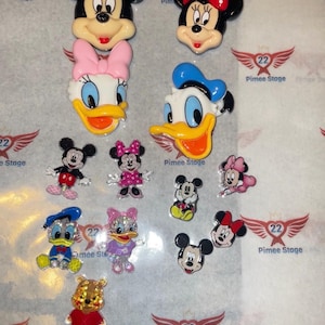 May include: A collection of Disney character-themed embellishments, including Mickey Mouse, Minnie Mouse, Donald Duck, Daisy Duck, and Winnie the Pooh. The embellishments are in various sizes and styles, with some featuring glitter and rhinestones.