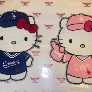 May include: Two Hello Kitty character patches, one in blue and one in pink, each wearing a baseball uniform. The blue patch has a blue cap with "LA" and a red bow. The pink patch has a pink cap with "LA" and a pink bow. Both have "Dodgers" on the jersey.