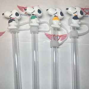 May include: Four clear plastic drinking straws with a white Snoopy character on top of each straw. The Snoopy characters are wearing different colored shirts: black, green, yellow, and black.