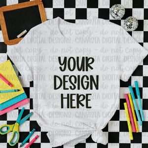 May include: White t-shirt with the text "YOUR DESIGN HERE" in a bold, black font. The shirt is styled with school supplies, including crayons, scissors, and a small chalkboard, set against a black and white checkered background.